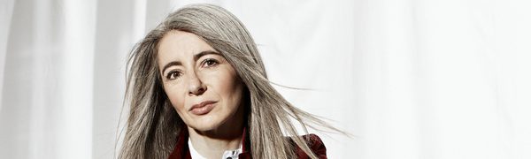 a woman with long grey hair leans against matte black percussion drums. She wears a maroon blazer, and looks at the camera with a serious expression.
