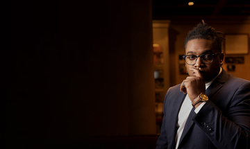 A man with glasses and black hair tied up, looks at the camera with his hand covering his mouth. He wears a suit, a large watch and a ring.