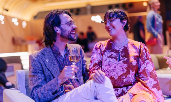 A man and a woman sit cosied up on a sofa laughing. The man holds a glass of wine while the woman rests her hand gently on his knee.