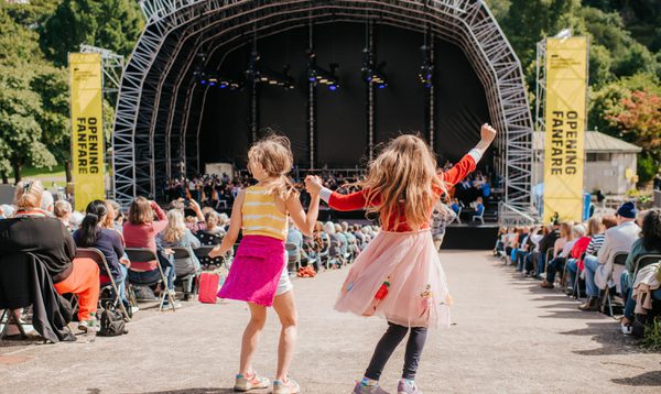 Two young girls earing bright outfits hold hands as they dance in the centre aisle of the Ross Bandstand seating. In the background, a large stage with yellow banners along the sides.