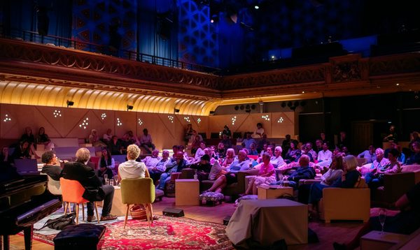 Three people sit on lounge chairs on a stage covered in rugs. An audience sitting on couches and lounge chairs watches and listens.
