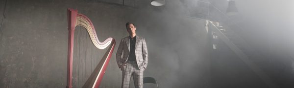 The harpist pictured standing next to his instrument in an atmospherically dark room, dressed in a black t-shirt and grey checked suit with his hands in his pockets.