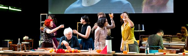 A group of people stand around a dining table with wine glasses underneath a screen playing the film Dogville