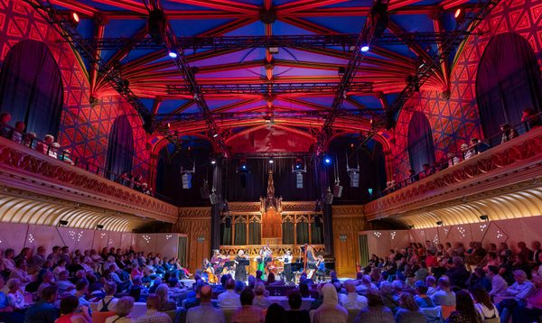 A band plays on stage, including a woman in a green dress playing the violin, and a harpist. In front of them an audience sits on couches. The ceiling is lit with vibrant colours.