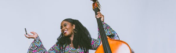 A smiling woman holds a double bass bow casually in her right hand as she looks up and away from the camera, holding up a double bass in her other hand.
