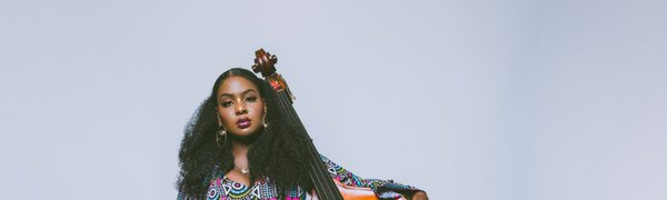A woman stands with her arm around a double bass, wearing a colourfully patterned mini dress and looking directly at the camera with a steady gaze.
