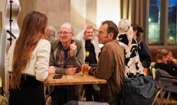 A woman with long brown hair talks to two men at a cafe table as they drink beer. They are all smiling.