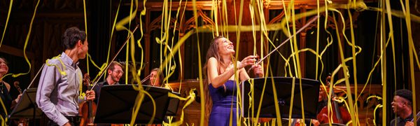 Nicola Benedetti, a white woman in a long purple dress, stands holding her violin with her head tipped back and smiling as yellow streamers float down with other musicians surrounding her
