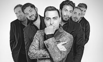 A man in a brocade suit jacket rests his chin in his palm gazing directly into the camera, with five men in dark suits standing gathered behind him.
