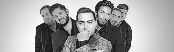 A man in a brocade suit jacket rests his chin in his palm gazing directly into the camera, with five men in dark suits standing gathered behind him.