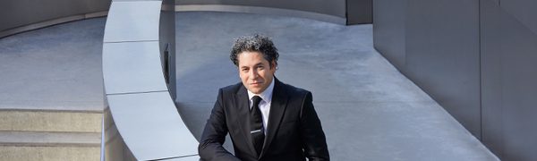 A man with short black curly hair, stands in a suit, leaning against a half wall, outside. The wall is curved, and the building is made from stainless steel.