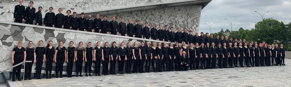 A choir of young people line up on slopes in front of a large grey building