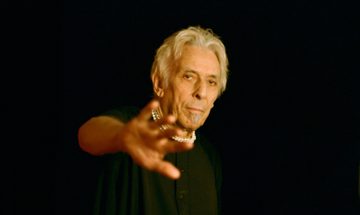 A man in a black t-shirt reaches one hand toward the camera. He stands in front of a black background.