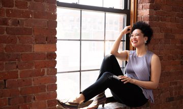 A smiling woman sits on a window ledge, with a brick wall behind her. She is wearing a purple silk shirt and her hair is a short afro