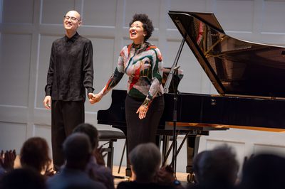 Two musicians stand in front of a grand piano, holding hands as they bow.