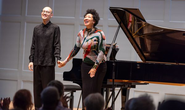 Two musicians stand in front of a grand piano, holding hands as they bow.
