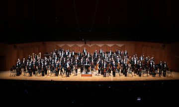 An orchestra stands on stage with their instruments, dressed in black tie