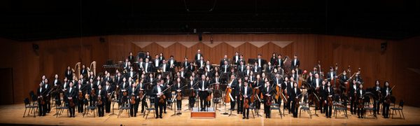 An orchestra stands on stage with their instruments, dressed in black tie