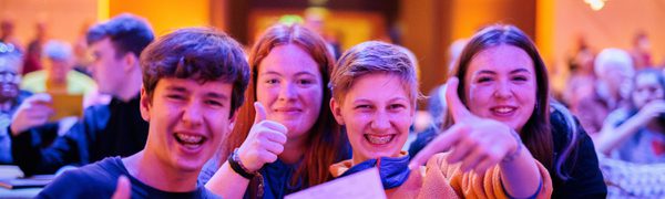 High school students sit on seats in a music venue. They grin holding up a ticket.
