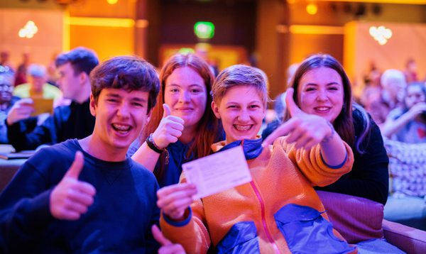 High school students sit on seats in a music venue. They grin holding up a ticket.