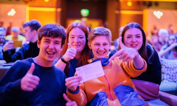 High school students sit on seats in a music venue. They grin holding up a ticket.