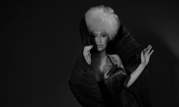 A woman with a big white afro looks straight ahead. She is wrapped in black silk fabric.