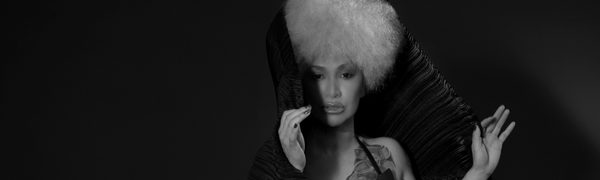 A woman with a big white afro looks straight ahead. She is wrapped in black silk fabric.