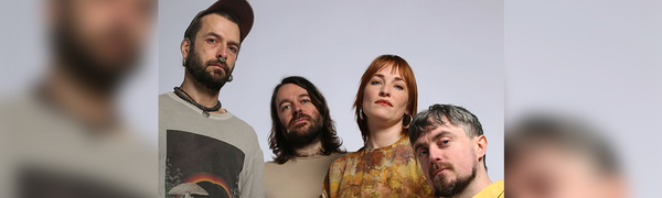 Three men and one woman look down towards the camera. They are dressed in neutral and yellow colours, and the background is grey. The woman's hair is orange, and one man wears a cap.