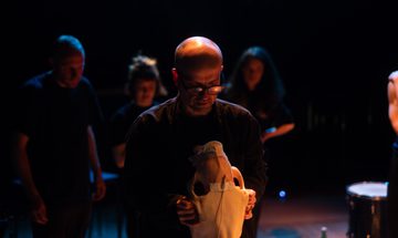 Matthew Herbert, wearing glasses and a black turtleneck, holds a horse head skeleton in two hands. He looks at it. Behind him at other musicians playing, also wearing black.
