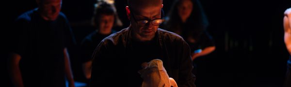 Matthew Herbert, wearing glasses and a black turtleneck, holds a horse head skeleton in two hands. He looks at it. Behind him at other musicians playing, also wearing black.