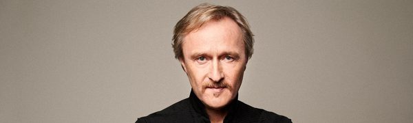 A blonde man with a moustache, dressed in a black button down top, stands in front of a beige photo backdrop.