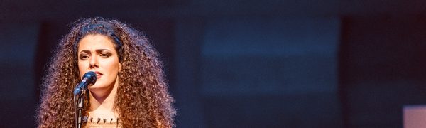 A woman with long curly hair sings into a microphone, looking intently forward and gesturing with her left hand.