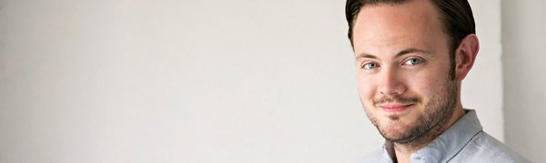 A man stands in front of a plain white background to the right of the frame. He's slightly smiling and wears a crisp blue button-down shirt.