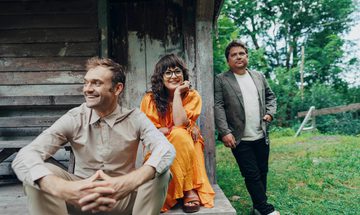Two men and a woman are outside a wooden building. The two men wear neutrals. The woman sits in between them and wears a bright orange dress