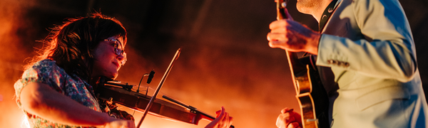 Two musicians face each other on an orange lit stage. The camera angle is looking up at them, as the woman on the left plays a fiddle and the man on the right plays an electric guitar.