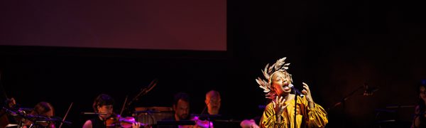 A woman sings in front of a microphone wearing a large gold dress and a gold head piece. Behind her is a band playing instruments.