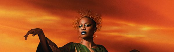 A woman standing against a fiery orange and yellow sky looks down at the camera. She's wearing a satin dress with puffed sleeves and holding her hands up and away from her.
