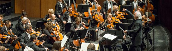 The strings section of the Deutsche Oper Berlin are being conducted by a man with white long hair to the left of the image.