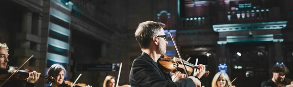 Six musicians play their fiddles, dressed in all black. In the background colourful lights emphasise the decorations of the inside of the building