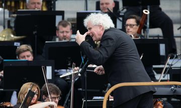 A white-haired man captured vigorously conducting an orchestra, turning to the site to gesture with his left hand. String and wind instrument players are visible behind music stands around him.