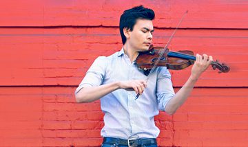 The violinist pictured in profile against a vibrant red background, wearing a blue shirt and jeans and playing his violin.