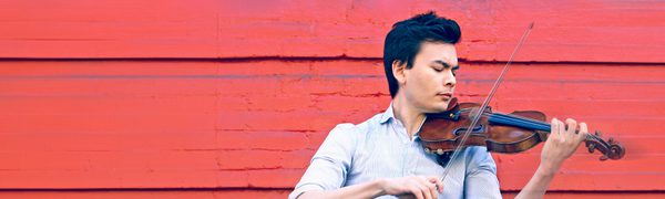 The violinist pictured in profile against a vibrant red background, wearing a blue shirt and jeans and playing his violin.
