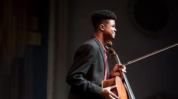 The cellist pictured in profile smiling onstage, holding his cello in front of him with the bow pointing towards the audience.