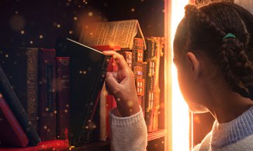 A child takes a book from a bookshelf and turns to look as a glowing portal appears to open in a gap between shelves.