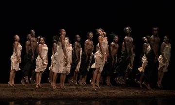 A group of dancers jump with their arms held straight against their legs in white dresses and black trousers above a floor covered in peat