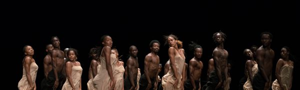 A group of dancers jump with their arms held straight against their legs in white dresses and black trousers above a floor covered in peat