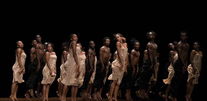 A group of dancers jump with their arms held straight against their legs in white dresses and black trousers above a floor covered in peat