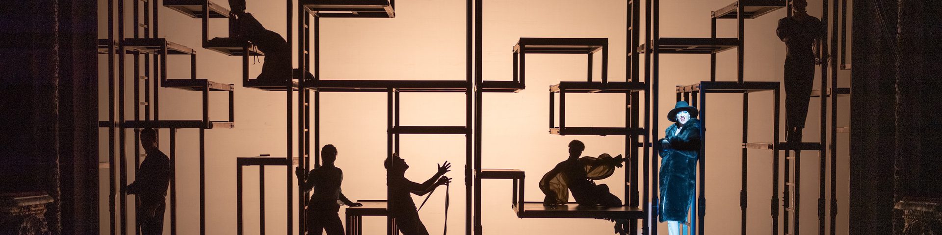 Silhouetted figures on complex scaffolding strike dramatic poses onstage, with a spotlight illuminating one singer in a fur coat, heels and black hat.