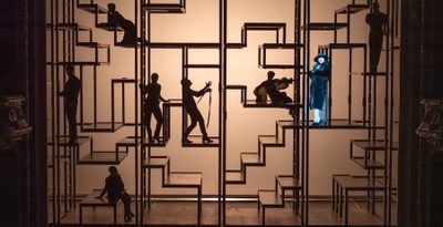 Silhouetted figures on complex scaffolding strike dramatic poses onstage, with a spotlight illuminating one singer in a fur coat, heels and black hat.