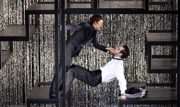 Two male actors dance, one holding a crab position as the other leans over him. They are on a metal structure.